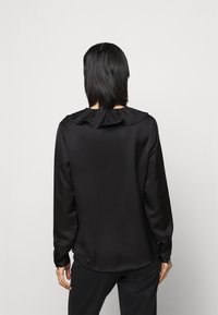 Svart satinblus med långa ärmar, volangkrage och rundad nederkant. Slät textur med minimalistisk design.