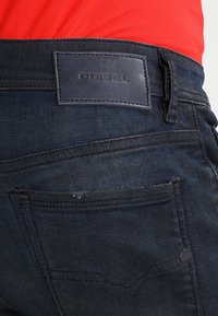 Jeans de mezclilla de indigo oscuro con una superficie texturizada, que cuentan con un parche de cuero negro con el logo de Diesel en la cinturilla y bolsillos traseros estándar.