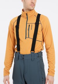 Oranje langwerpig T-shirt met ritssluiting en grijze stiksel, gecombineerd met blauwe broeken met zwarte bretels en meerdere zakken.