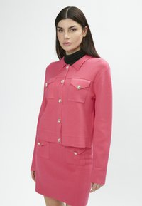 Conjunto de cárdigan y falda en una tela texturizada. Presenta cuatro bolsillos y botones plateados brillantes. Diseño en capas con cuello de solapa.