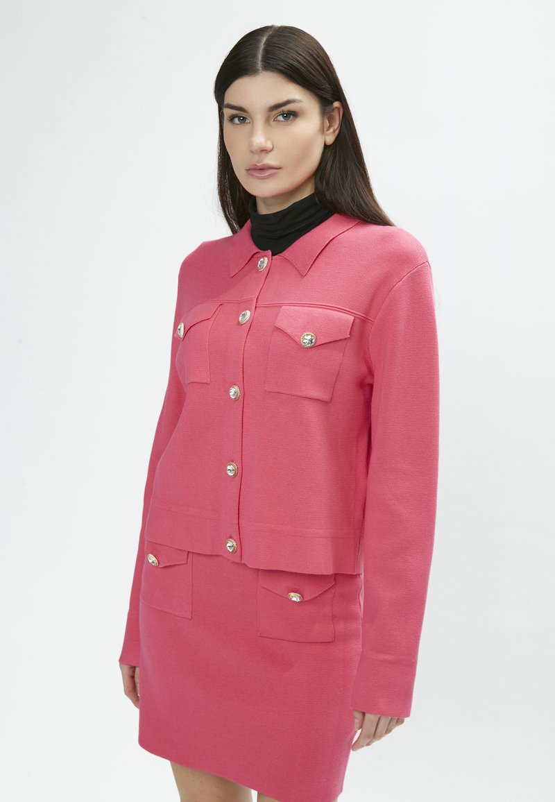 Conjunto de cárdigan y falda en una tela texturizada. Presenta cuatro bolsillos y botones plateados brillantes. Diseño en capas con cuello de solapa.