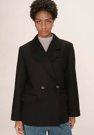 Femme aux cheveux courts et bouclés, portant un blazer croisé noir sur un haut gris et un jean bleu, se tenant contre un fond clair et uni.