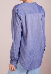 Camisa de manga larga de color azul claro hecha de tejido suave. Presenta un espolón en la parte trasera, puños de manga con botones y un dobladillo ligeramente curvado.