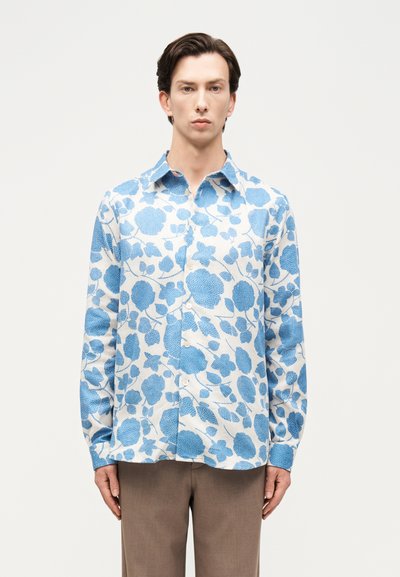 Joven con una camisa blanca de botones con un patrón floral azul y pantalones marrones, de pie frente a un fondo blanco liso.