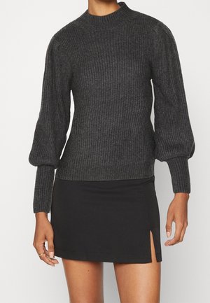 Vrouw draagt een donkergrijze geribbelde gebreide trui met ballonmouwen en een zwarte minirok met een voorsplit.