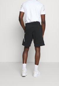 Černé bavlněné šortky s elastickým pasem, postranními kapsami a bílým logem na levé noze. Viditelné bílé tenisky a ponožky do půli lýtek.