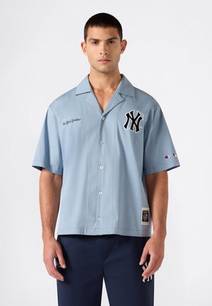 Mann trägt ein hellblaues New York Yankees Hemd mit kurzen Ärmeln und dunkler Hose vor einem einfarbigen weißen Hintergrund.