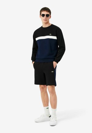 Uomo che indossa una felpa nera, bianca e blu navy con pantaloncini neri abbinati, calzini bianchi, scarpe da ginnastica bianche e occhiali da sole scuri, in piedi con le mani in tasca.