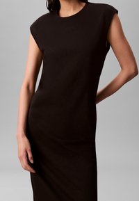Robe noire sans manches avec épaules rembourrées portée par une personne avec un bras plié derrière et l'autre pendant.