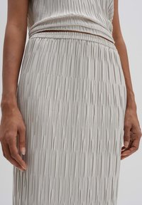 Plooienrok in lichtgrijs met een elastische tailleband, gladde textuur en verticale lijnen. Close-up toont detail van de stof en handpositie.