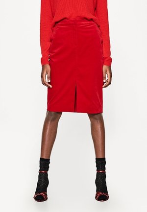 Mujer que lleva una falda roja hasta la rodilla con abertura frontal, jersey rojo, calcetines negros con brillo y tacones rojos abiertos, de pie sobre un fondo blanco.
