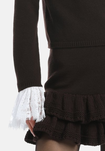 Pull en laine marron avec des poignets évasés en dentelle blanche et un bas à volants. La texture est douce, avec un design superposé et confortable.