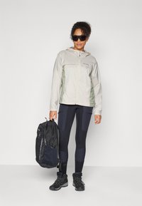 Kevyt, beige ja vihreä takki vetoketjulla, yhdistettynä mustiin legginsseihin ja mustaan reppuun. Asusteina tummat aurinkolasit.