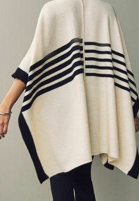 Poncho crema con rayas horizontales negras, que presenta una textura suave y un corte holgado, con apertura frontal y mangas cortas. Los bordes tienen un ribete oscuro.