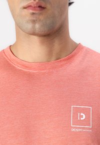Koralo raudonos T-shirt marškiniai iš minkštos medvilnės medžiagos su apvaliu kaklu. Ant priekio kairėje pusėje yra baltas kvadratinis logotipas ir paprastas tekstas. Nepriekaištinga tekstūra.