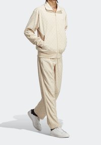 Béžový teplákový komplet s jemným vzorem listů, zapínací bunda, elastické manžety a odpovídající kalhoty. V kombinaci s bílými teniskami.