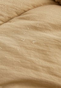 Bēša audums ar strukturētu virsmu, kas satur niansētu spīdumu un nelielas iestrādes visā virsmā. Mīksts un nedaudz burzīts izskats.