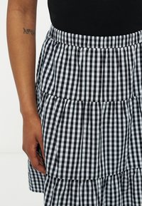 Ασπρόμαυρη φούστα με καρώ μοτίβο σε στρώσεις, φορεμένη με μαύρο τοπ, με το αριστερό χέρι να φαίνεται και ένα τατουάζ με γραμμικό σχέδιο κοντά στον αγκώνα.