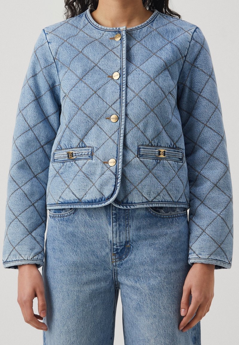Personne portant une veste en jean bleu clair matelassée avec des boutons dorés et un jean assorti ; seuls le torse et les bras sont visibles.