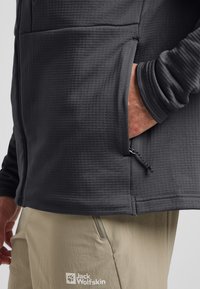 Veste grise à manches longues avec un tissu texturé et une poche zippée. Pantalon beige avec un logo sur la cuisse. Accent sur les vêtements de plein air.