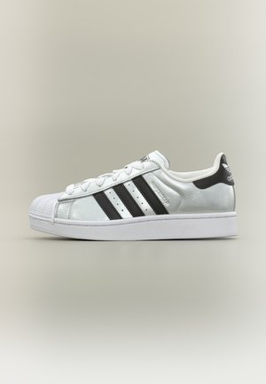 Baskets blanches Adidas Superstar avec bandes noires et languette au talon, embout strié et semelle blanche sur fond neutre.