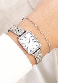 Montre en métal argenté avec un cadran rectangulaire, un cadran blanc, des chiffres romains et un bracelet tissé. Portée avec un bracelet en chaîne délicat en argent.