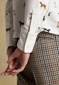 Ruky prekrížené za chrbtom, oblečené v bielej košeli s potlačou psíkov, s manžetami na zápästí, cez multikolorové kárované nohavice s gombíkovou kapsou.