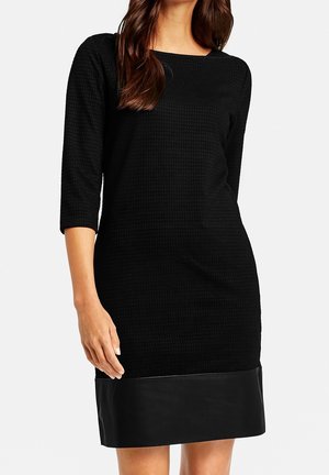 Woman wearing a black textured knee-length dress with three-quarter sleeves and a smooth leather hem.