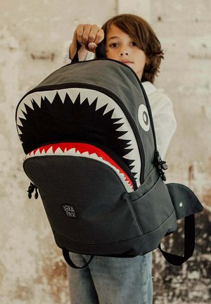 Backpack in dark gray with shark-mouth design. Features white teeth and red accents. Adjustable straps and front pocket with zipper.