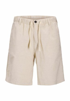 Garçon debout portant une chemise à manches courtes à carreaux noir et blanc, un short beige, des chaussettes blanches et des baskets blanches sur un fond uni.