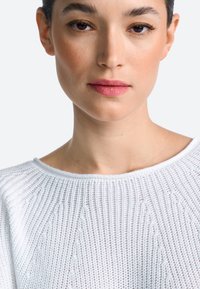 Biely pletený sveter s rebrovanou textúrou a širokým výstrihom. Modelka má neutrálny výraz, jemný make-up a tmavé vlasy.