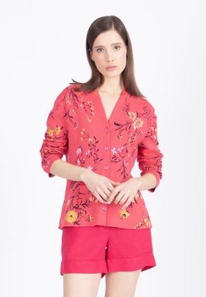Mujer con un cárdigan de botones de flores rosas y pantalones cortos rosas lisos a juego, de pie contra un fondo blanco liso.
