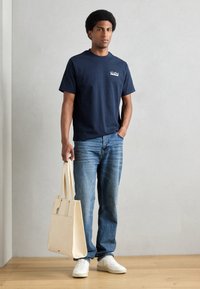 Marine blå bomull t-skjorte med en liten logo, brukt sammen med lyseblå jeans. En beige tote bag i den ene hånden og hvite joggesko fullfører antrekket.