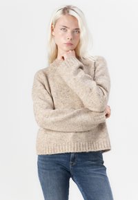 Beige gestrickter Pullover mit Rundhalsausschnitt und gerippten Bündchen. Kombiniert mit blauen Jeans. Das strukturierte Gewebe zeigt Einschlüsse in dunklerer Farbe.