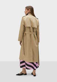 Beige trench coat with embossed logo pattern, belted waist, shoulder epaulettes, and a back vent, paired with a pink and black striped skirt.
