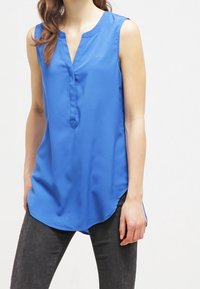 Blusa senza maniche di un blu profondo con scollatura a V, texture liscia e orlo curvo. Presenta una pattina frontale e un design minimalista.