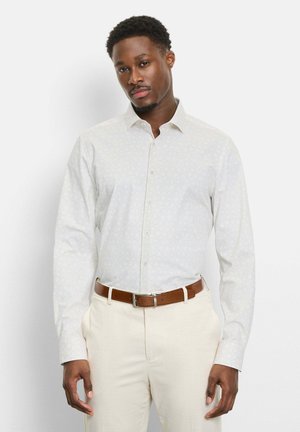 Mann trägt ein weißes, gemustertes Langarmhemd, in cremefarbene Hosen mit braunem Gürtel gesteckt, steht vor einem schlichten weißen Hintergrund.