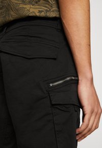 Pantaloni cargo negri, cu multiple buzunare, inclusiv un buzunar lateral cu fermoar, și un finisaj din țesătură texturată.