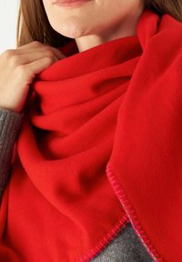 Rode wollen sjaal om de nek gedrapeerd, met een gestructureerd oppervlak en contrasterende roze stiksels langs de rand, gelaagd over een grijze kledingstuk.