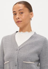 Γκρίζα πλεχτή ζακέτα με σχέδιο σε V, δύο μπροστινές τσέπες με κρεμ προφίλ και κλείσιμο με κουμπί. Λείο υλικό και ελαφριά υφή.