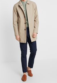 Beige wasserdichter Mantel mit Knopfverschluss und Kragen. Getragen über einem grünen Hemd und dunkelblauen Hosen, kombiniert mit braunen Slippern.