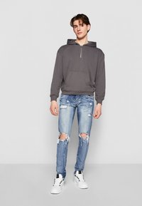 Haut à capuche zippé gris avec poche avant, associé à un jean slim déchiré bleu clair. Le mannequin porte des baskets blanches. Arrière-plan simple.