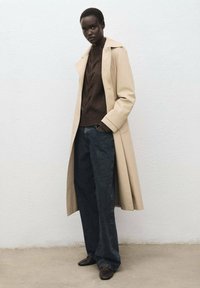 Cappotto beige, maglione a trecce marrone scuro, jeans larghi scuri e scarpe marroni. Linee pulite, texture sovrapposte e design minimal.