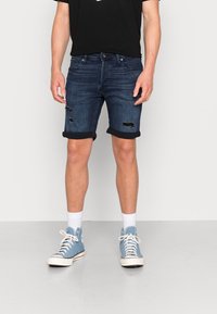 Tummansiniset farkkushortsit, joissa on käänteet ja kulutetut yksityiskohdat. Yhdistetty mustan t-paidan ja sinisten korkeaotsisten tennareiden kanssa.