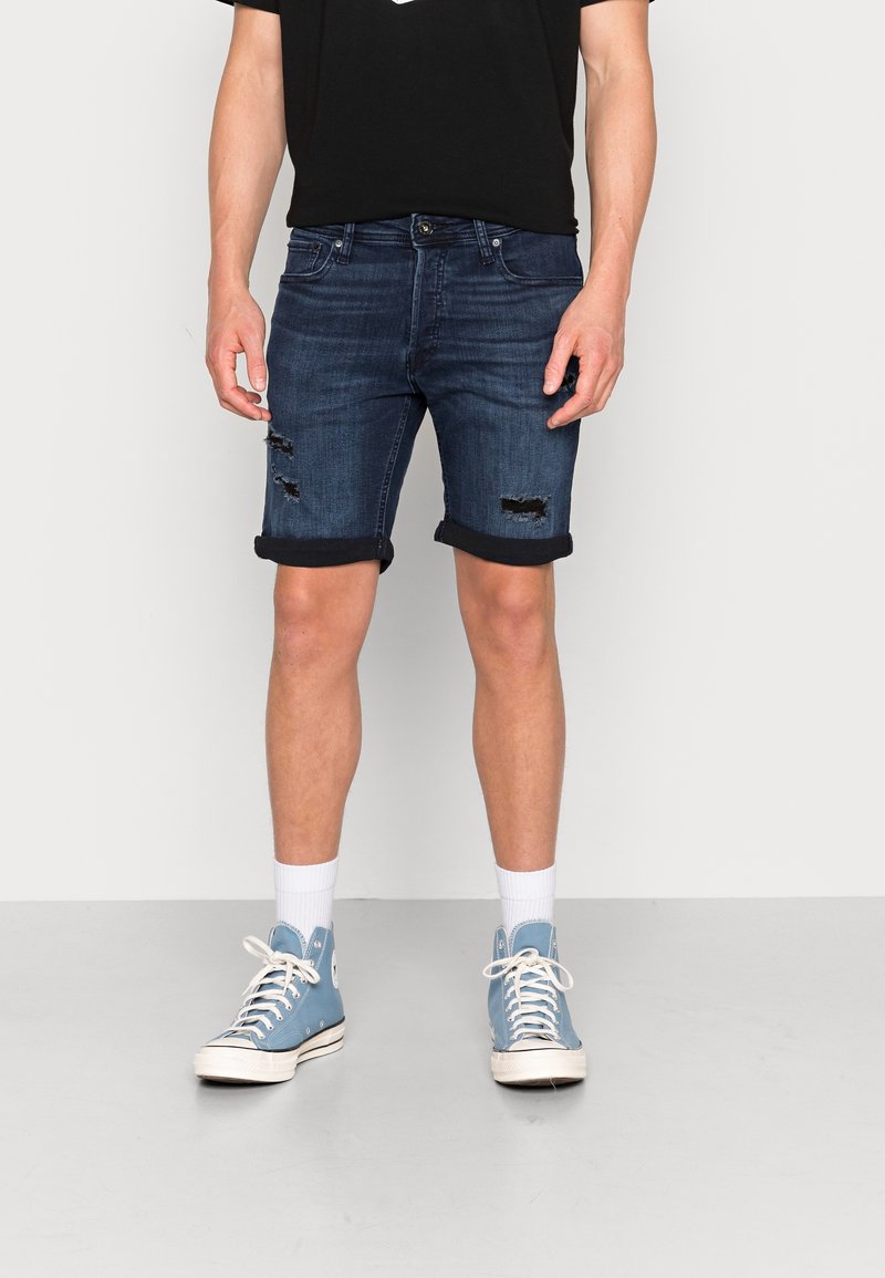 Tummansiniset farkkushortsit, joissa on käänteet ja kulutetut yksityiskohdat. Yhdistetty mustan t-paidan ja sinisten korkeaotsisten tennareiden kanssa.