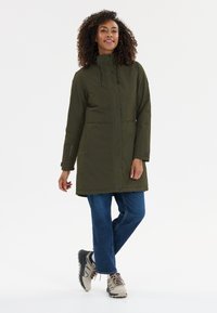 Olivgrön, vattenavvisande lång kappa med hög krage, framfickor och justerbar dragsnodd. Kombinerad med blå jeans och lätta sneakers.