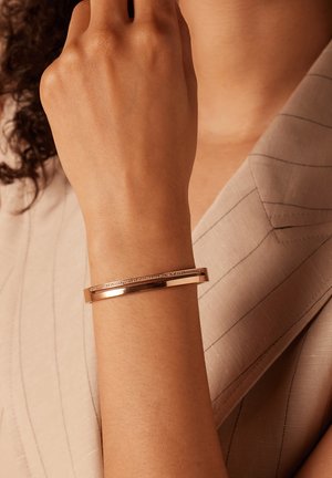 Bracelet en métal rose doré avec une surface lisse et une rangée de petits cristaux transparents le long du bord supérieur. Porté au poignet.