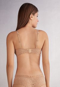 Sostenedor de encaje nude con tirantes ajustables y cierre de gancho. Presenta un intrincado patrón floral a lo largo de la banda y bordes en puntilla.