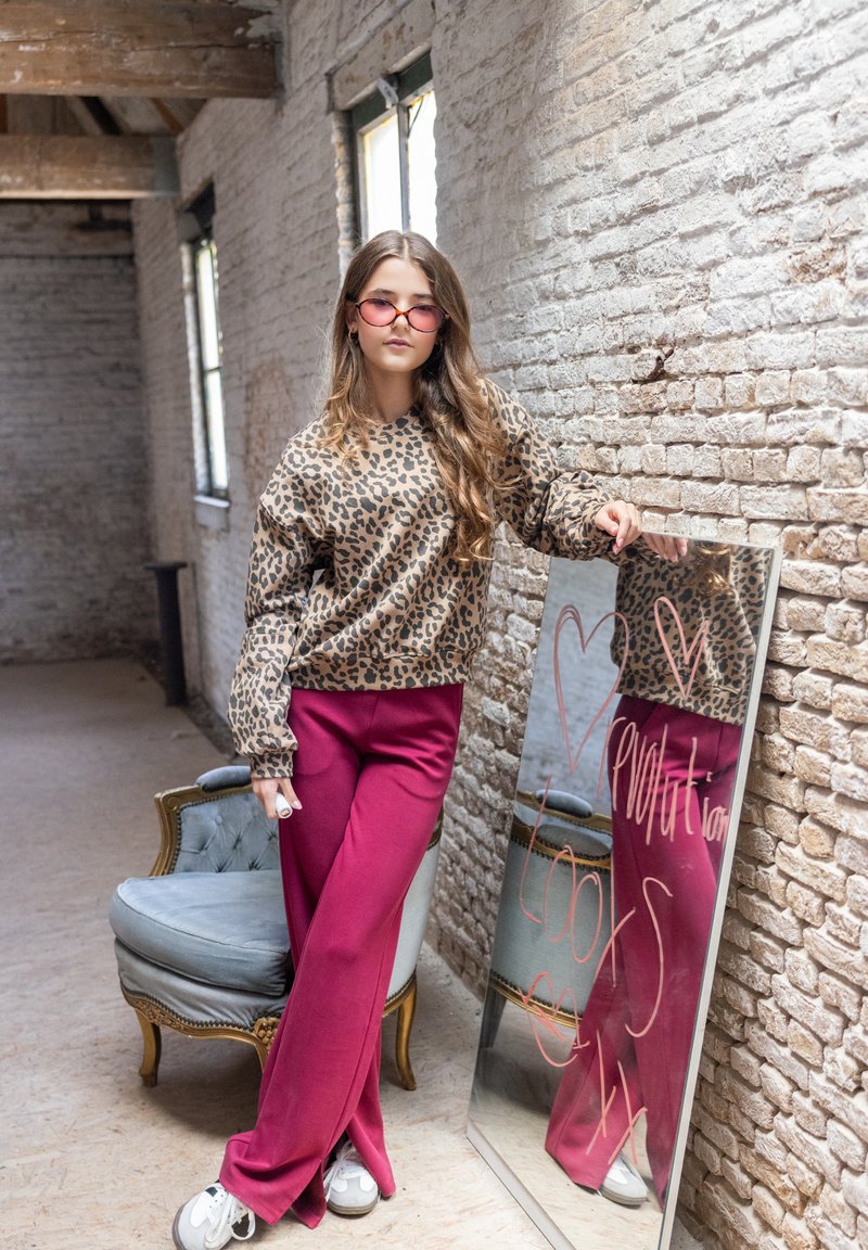 Luipaardprint sweatshirt, roze wijde broek, witte sneakers; staande bij een spiegel in een rustieke setting met zichtbare bakstenen muren.