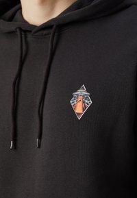 Felpa con cappuccio nera in cotone, con un piccolo e colorato emblema grafico raffigurante un vulcano all'interno di una forma a diamante sul petto.